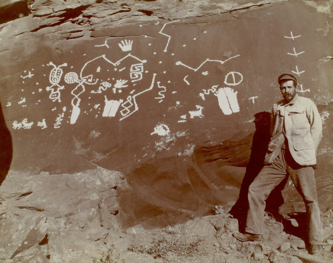 Historic photograph of a man standing beside prehistoric rock art painted on a stone wall.